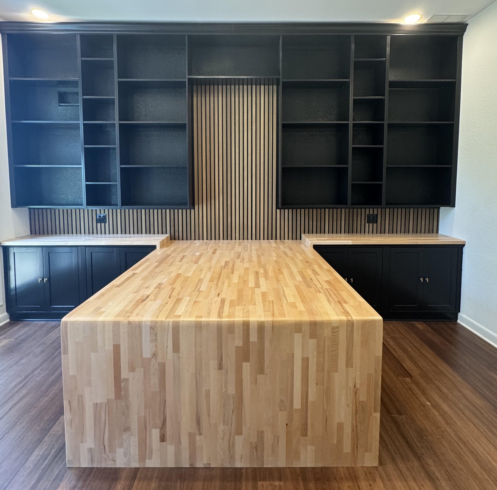 Floor-to-ceiling black built-in office wall, symmetrical either side of a fluted oak slat panel, with a maple butcher-block desk running out into the room