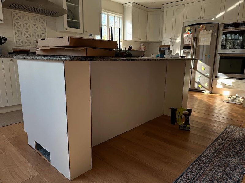Kitchen island under construction with cream cabinetry and a granite top being fitted over hardwood floors