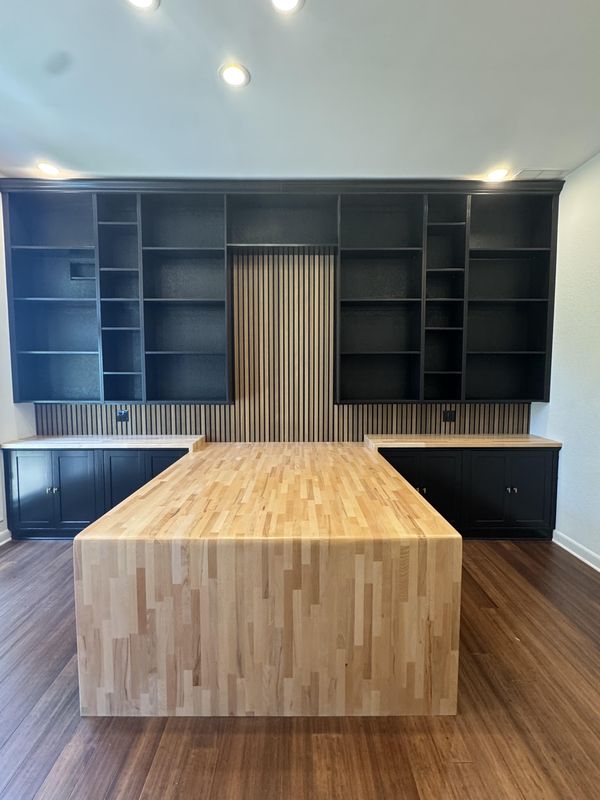 Floor-to-ceiling black built-in office wall, symmetrical either side of a fluted oak slat panel, with a maple butcher-block desk running out into the room